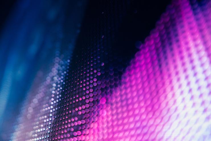 Eine abstrakte, unscharfe Nahaufnahme zeigt lila und blaue Lichtpunkte auf einer strukturierten Oberfläche, die ein futuristisches, digitales oder technologisches Gefühl vermittelt.