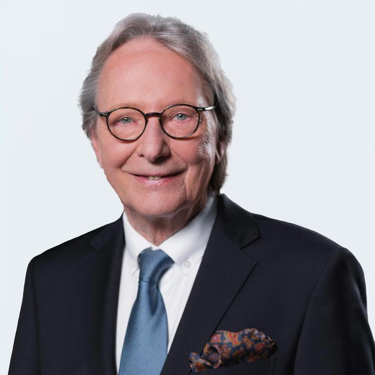 Dr. Jürgen Apel, ein älterer Herr mit Brille und grauen Haaren, trägt einen dunklen Anzug, blauen Schlips und ein farbiges Einstecktuch, während er freundlich in die Kamera lächelt.